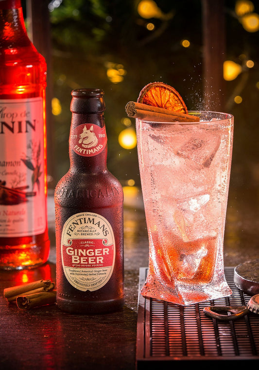 A bottle of Fentimans Ginger Beer beside a highball drink garnished with orange and cinnamon with a bottle of Monin in the background